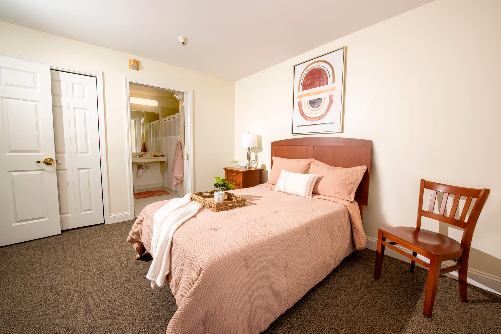 A cozy bedroom with a neatly made bed featuring a light pink bedspread and pillows. A wooden headboard is mounted against a cream-colored wall with an abstract framed artwork above it. A wooden nightstand with a lamp and small plant sits next to the bed. A wooden chair is placed against the opposite wall. An open door reveals a bathroom with a shower curtain, sink, and mirror. The room has beige carpet flooring and white doors.