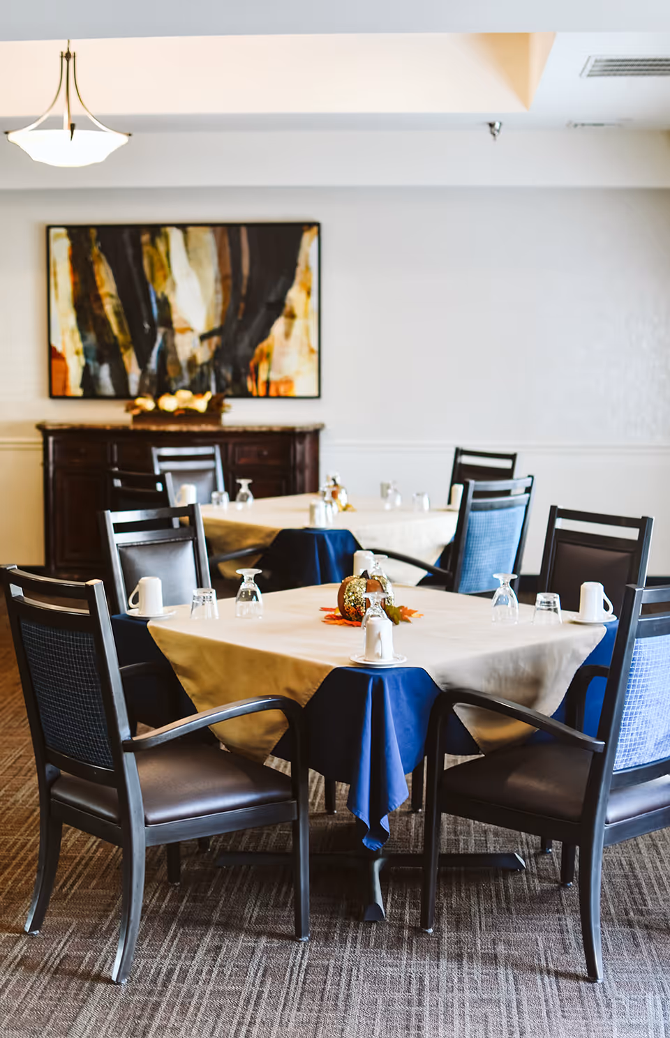 A dining room with tables covered in beige and blue tablecloths, set with upside-down glasses and white coffee mugs. Dark wooden chairs with armrests surround the tables. A sideboard with decorative items and a large abstract painting hang on the wall in the background.