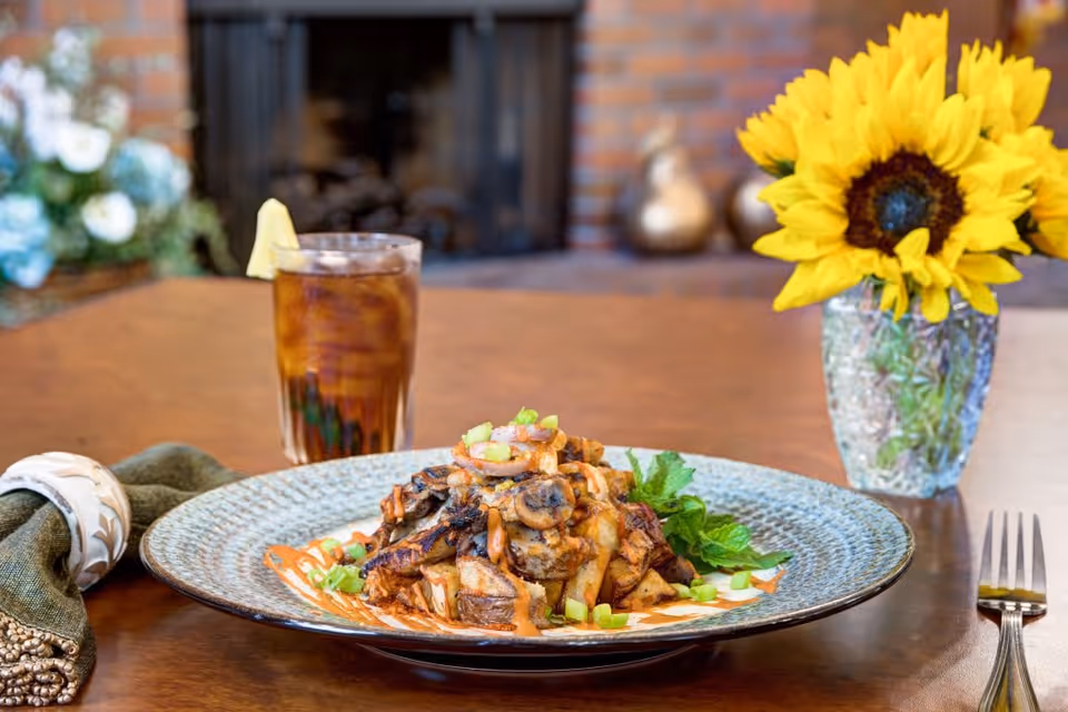A plate of cooked mushrooms and potatoes garnished with green onions and sauce on a blue patterned plate, placed on a wooden table. Next to the plate is a glass of iced tea with a lemon wedge, a green cloth napkin with a white napkin ring, a fork, and a vase with a bright yellow sunflower. The background shows a blurred fireplace and some decorative items.