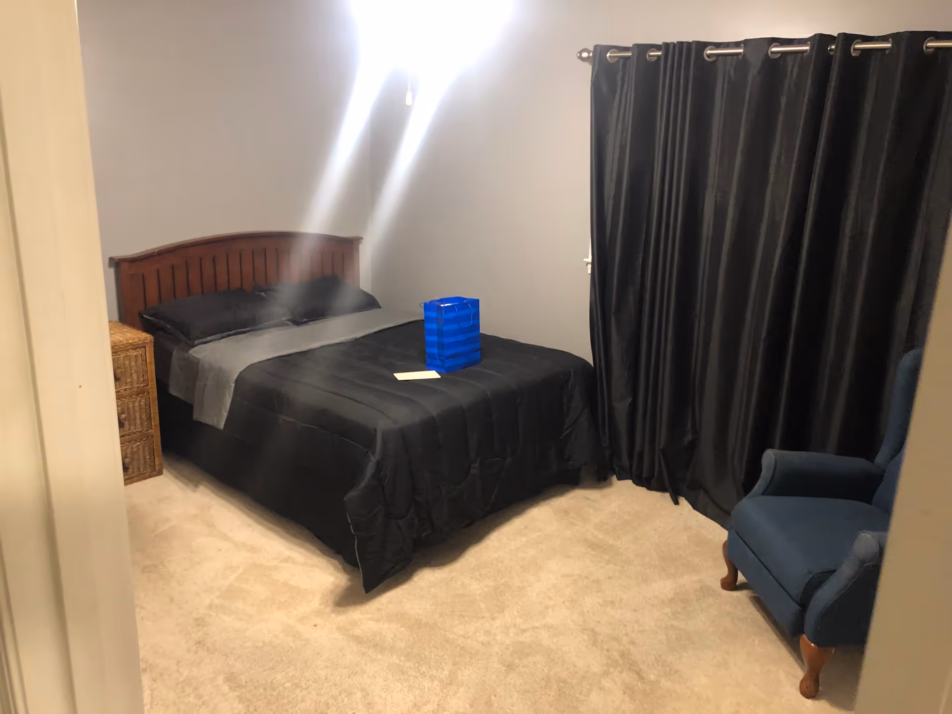 Simple bedroom with a bed dressed in black linens, a wicker nightstand, dark curtains, and a blue armchair.