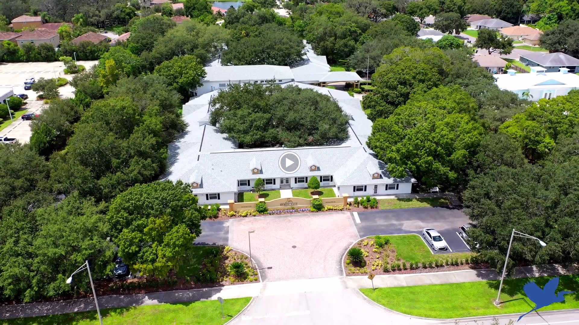 Aerial view of a senior living facility named Heron East surrounded by dense green trees and landscaping. The building has a light gray roof and white exterior walls, with a driveway and parking area in front. There are several cars parked and a well-maintained lawn and garden areas around the property.