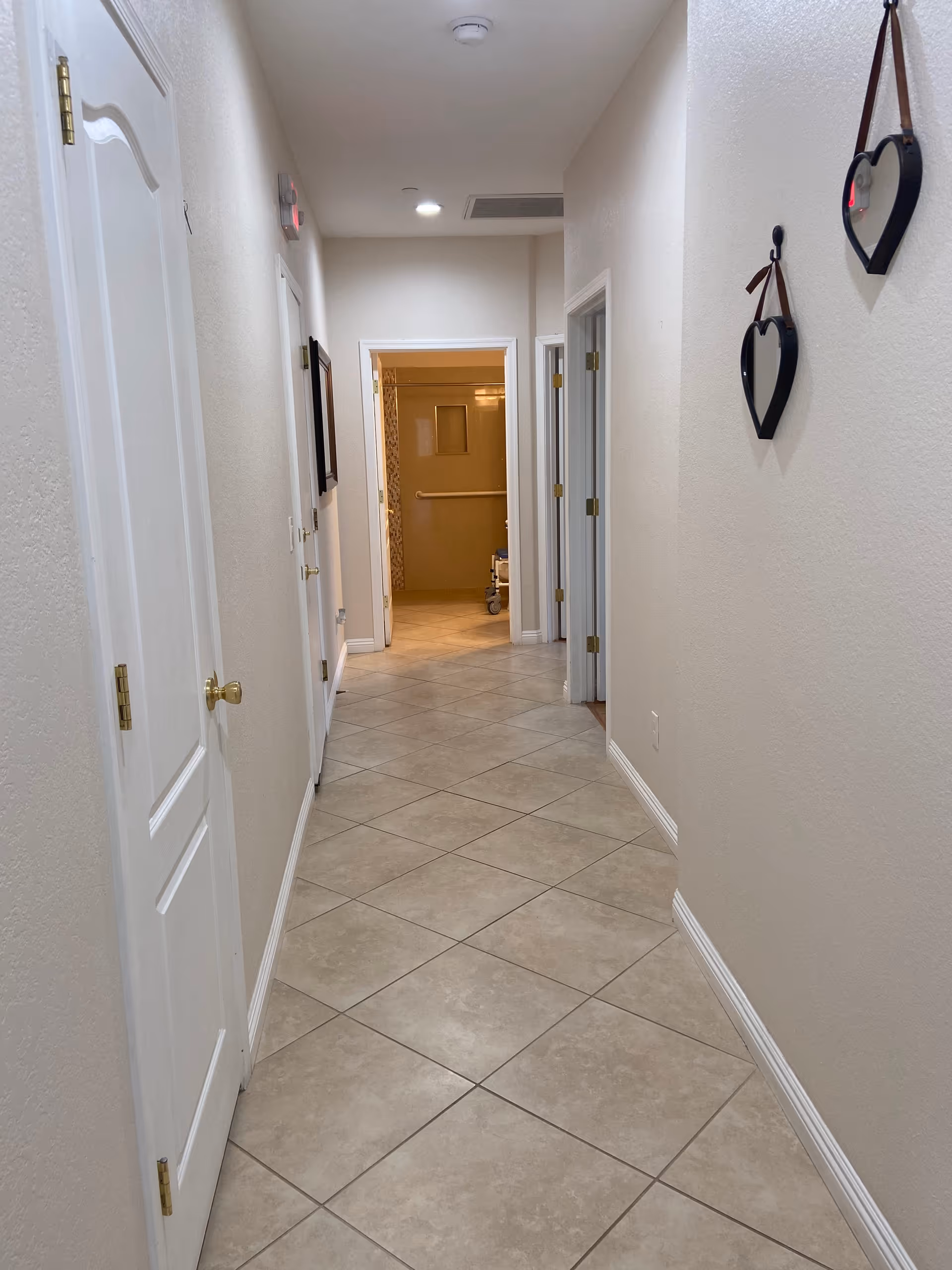 A clean, well-lit hallway with beige tiled flooring and cream-colored walls. There are several white doors along the hallway, and two heart-shaped wall decorations hanging on the right wall. At the end of the hallway, there is an open doorway leading to another room with a grab bar and a walker visible.