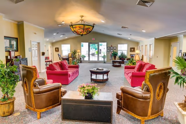 Spacious, well-lit senior living lounge with red sofas, leather armchairs, coffee tables, plants, and a stained-glass chandelier.