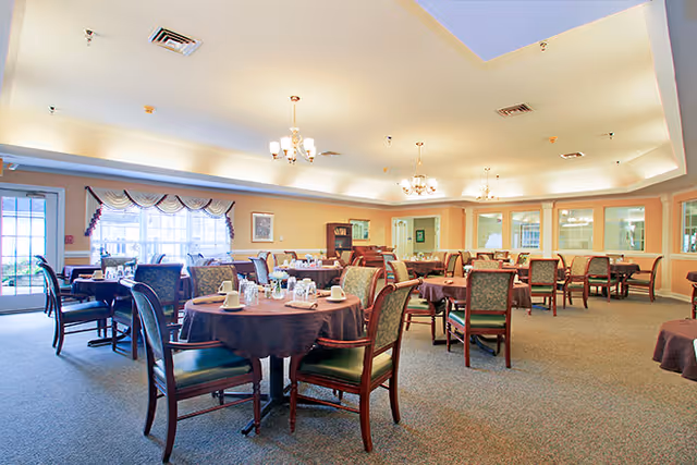 A spacious dining room with multiple round tables covered with dark tablecloths, each set with cups, saucers, and utensils. The room has large windows with valances, chandeliers hanging from the ceiling, and upholstered chairs around the tables. The walls are painted in warm tones and there is carpeted flooring.