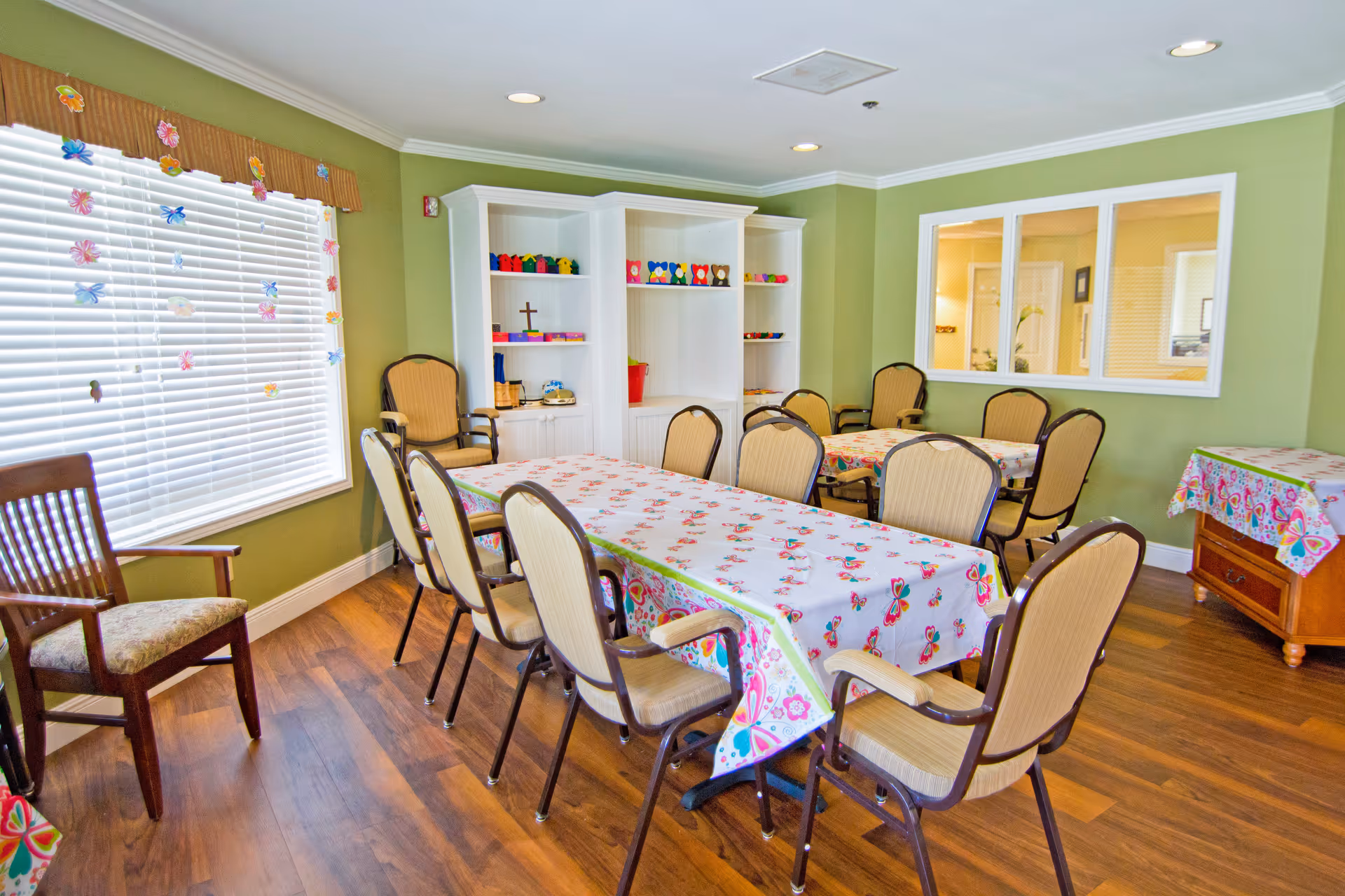 A bright and cheerful dining room with light green walls and wood flooring. There are two tables covered with colorful butterfly-themed tablecloths, surrounded by beige cushioned chairs with dark frames. A large window with white blinds and butterfly decorations lets in natural light. White shelving units with small colorful decorative items and a cross are against the wall. A wooden side table with a matching butterfly tablecloth is in the corner, and a large interior window looks into another room.