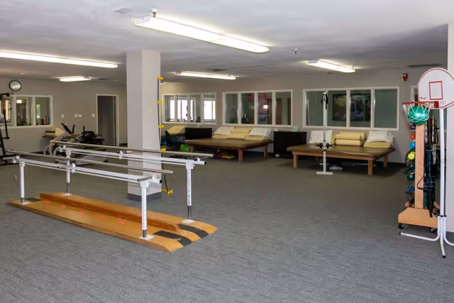 A spacious rehabilitation room with parallel bars for walking exercises in the foreground, several padded therapy tables along the back wall, exercise equipment including a stationary bike and a basketball hoop with a ball rack on the right side, and large windows looking into adjacent rooms.