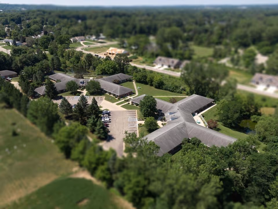 Aerial view of a single-story senior living campus with interconnected buildings, parking lot, and surrounding trees and fields.