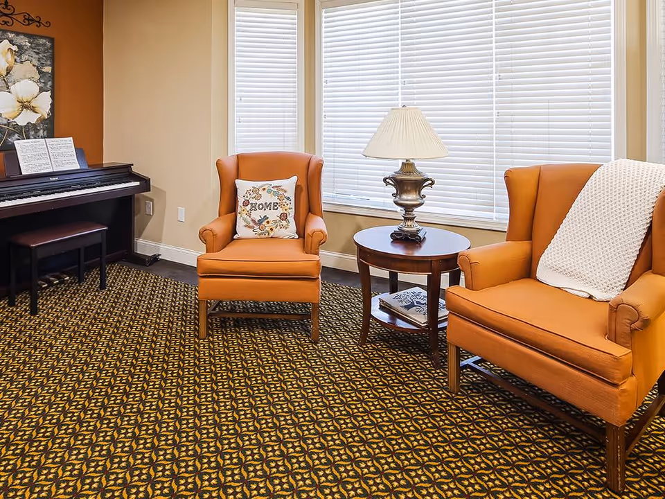 Cozy seating area with two orange armchairs, a round side table with a lamp, and a piano against the wall.
