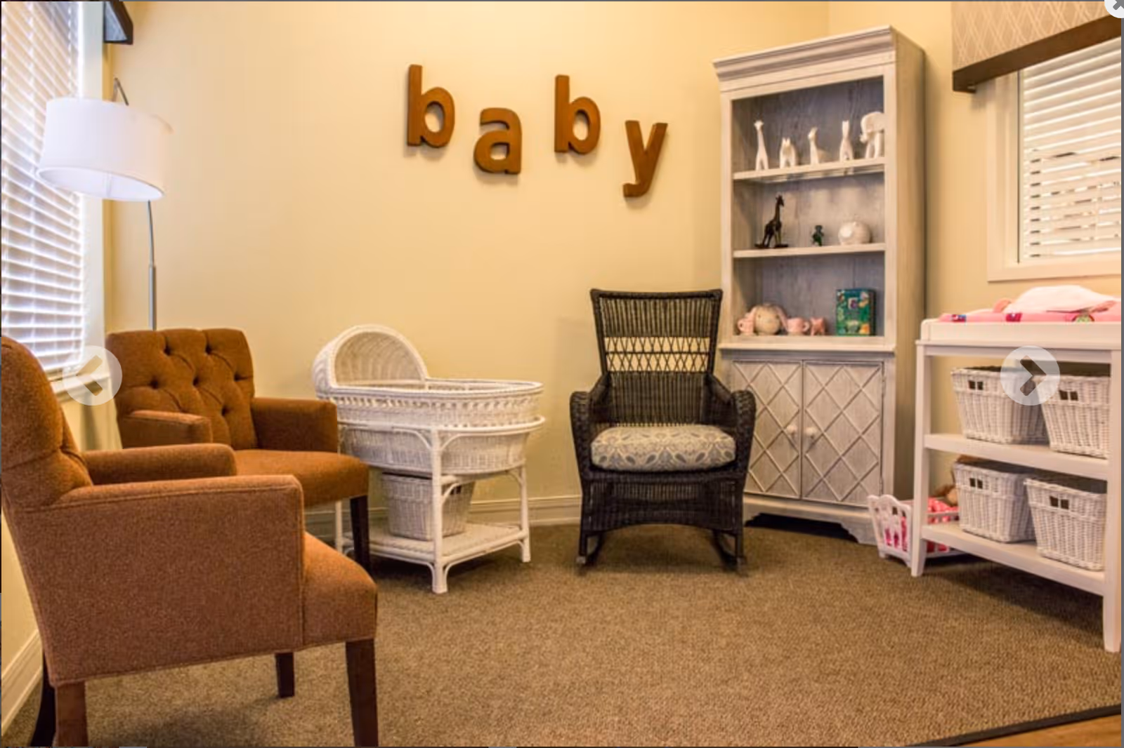 Cozy nursery-style room with chairs, a wicker bassinet, shelving, and wooden letters spelling "baby" on the wall.