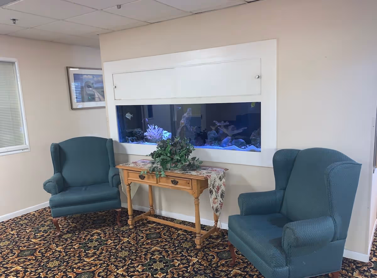 A cozy sitting area with two teal upholstered wingback chairs facing a wooden table with a floral runner and a green potted plant. Behind the table is a built-in wall aquarium with coral and fish. The room has a patterned carpet and light-colored walls with a framed picture and a window with blinds.
