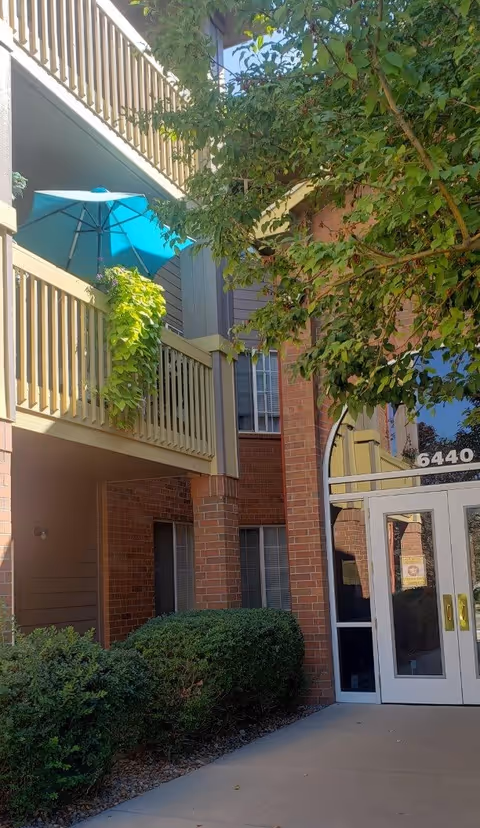 Exterior view of a senior living facility named Springwood showing a brick building entrance with glass double doors, a balcony with a blue umbrella and green plants, and surrounding bushes and trees.
