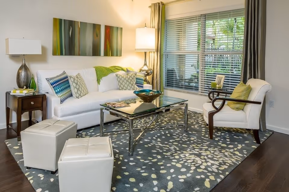 Bright living room with a white sofa, armchair, glass coffee table, patterned rug and a large window with blinds.