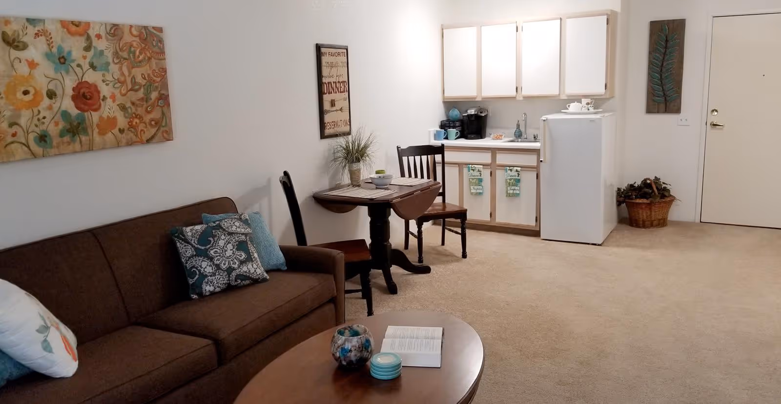 Cozy studio-style living area with a brown sofa, coffee table, small dining table and chairs, and a kitchenette with a mini-fridge.