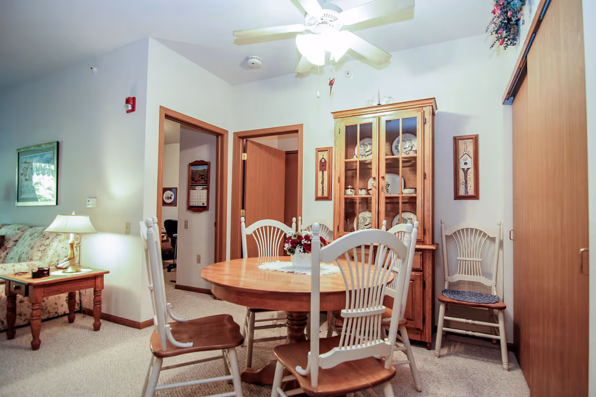 A cozy dining area in a senior living apartment featuring a round wooden table surrounded by five wooden chairs with white backs and brown seats. Behind the table is a wooden china cabinet displaying plates and teapots. The room has light-colored walls, a ceiling fan with a light, and two doorways leading to other rooms. To the left, part of a floral-patterned couch and a wooden side table with a lamp are visible.