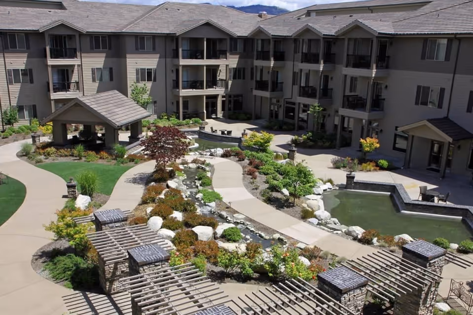 Outdoor courtyard area of a senior living facility featuring a landscaped garden with a small pond, walking paths, shrubs, and trees. The courtyard is surrounded by a multi-story building with balconies and windows. There are pergolas and seating areas around the garden.