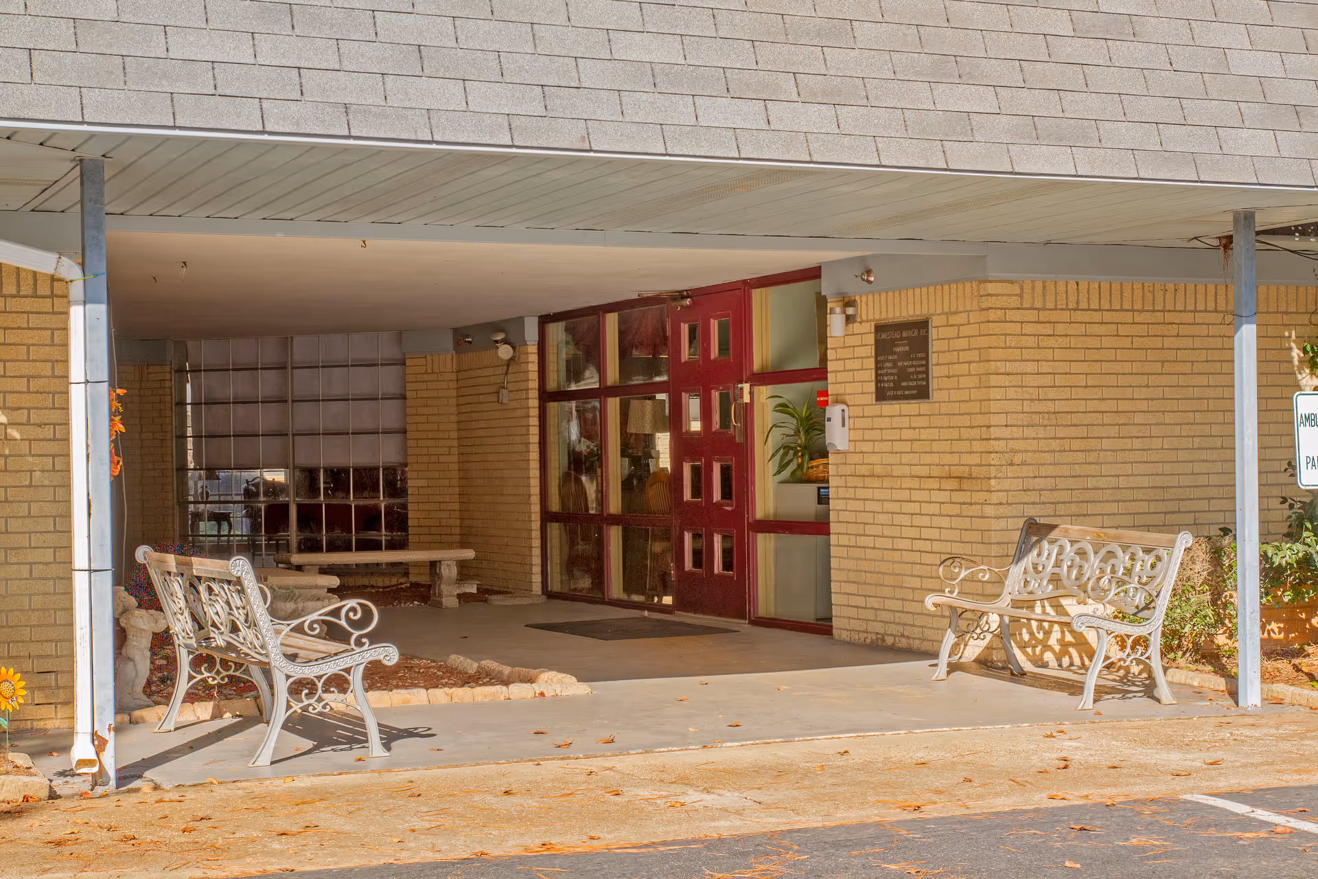 Entrance area of a building with a covered walkway, two white decorative metal benches on either side, a red door with glass panels, and beige brick walls. There is a small garden bed with a stone bench and some plants visible inside the covered area.