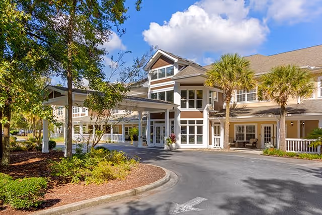 Exterior view of a senior living facility building with a covered entrance, palm trees, and landscaped greenery under a blue sky with some clouds.