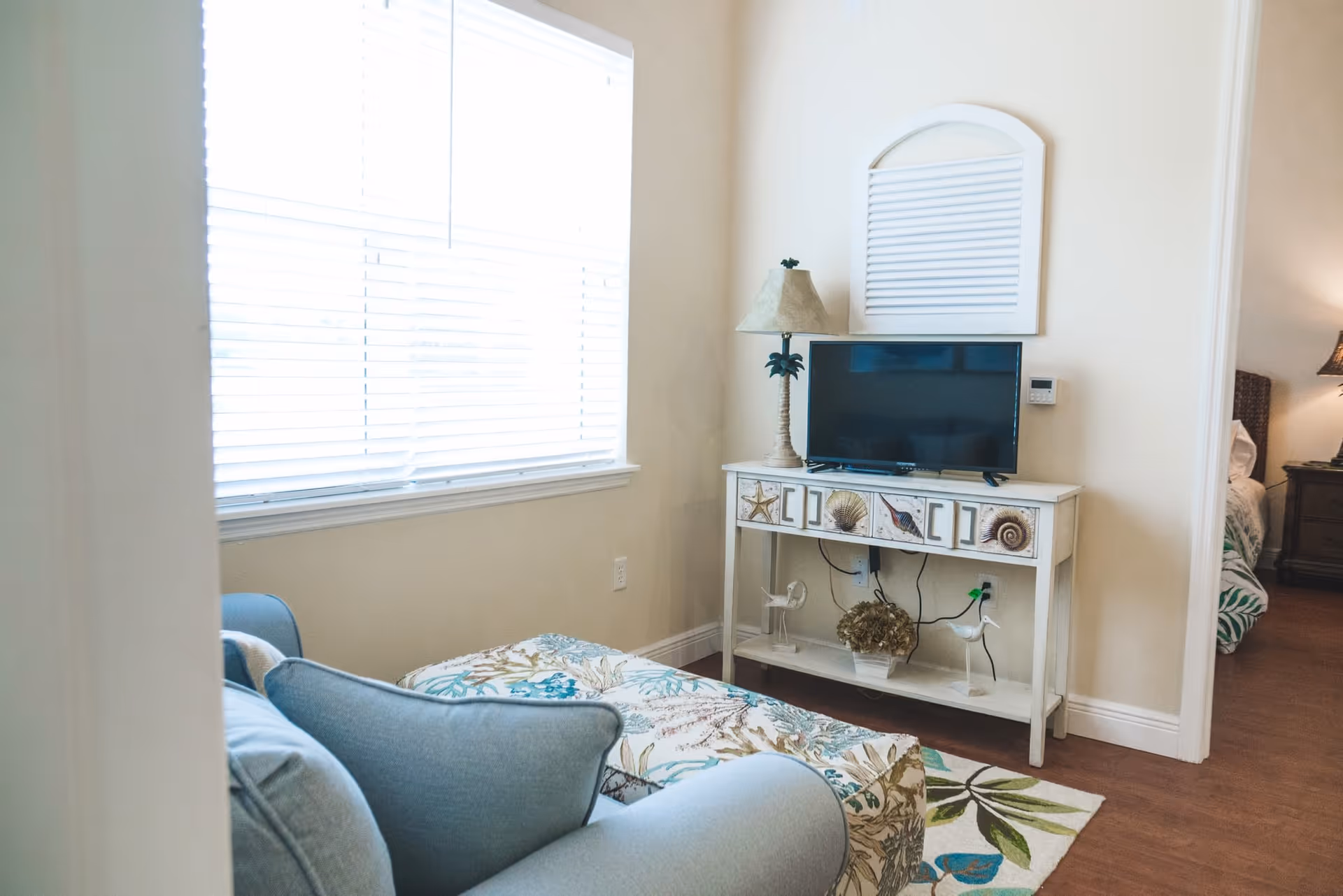 A cozy living room area with a light blue armchair and a floral-patterned ottoman. A white console table with seashell decorations holds a flat-screen TV and a lamp with a palm tree base. There is a large window with white blinds letting in natural light, and an adjacent room with a bed and nightstand is partially visible.