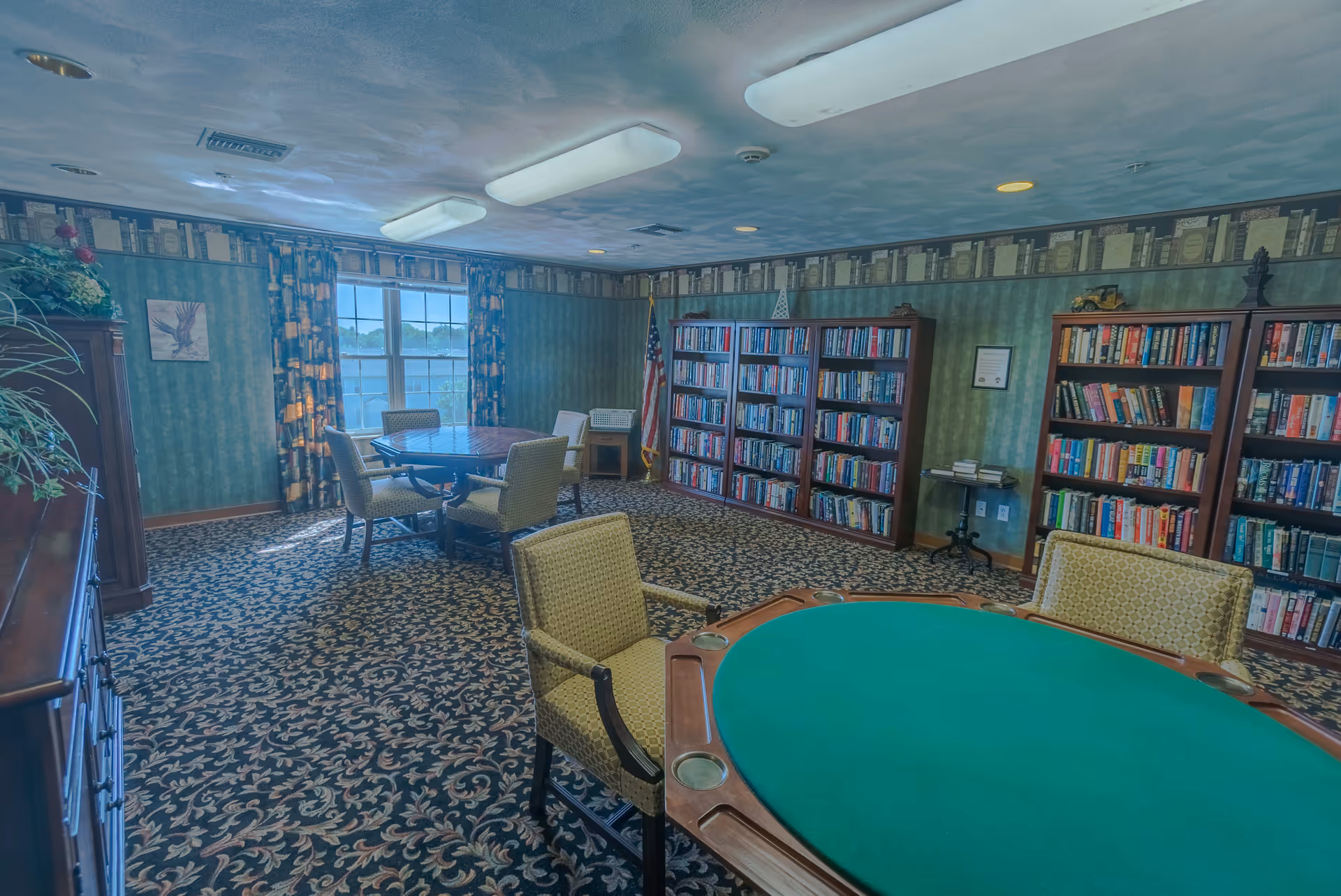 A cozy interior room with patterned carpet and wallpaper, featuring multiple bookshelves filled with books along the walls. There are two round tables with chairs around them, one table has a green felt surface, possibly for card games. A window with patterned curtains lets in natural light, and an American flag stands in the corner.