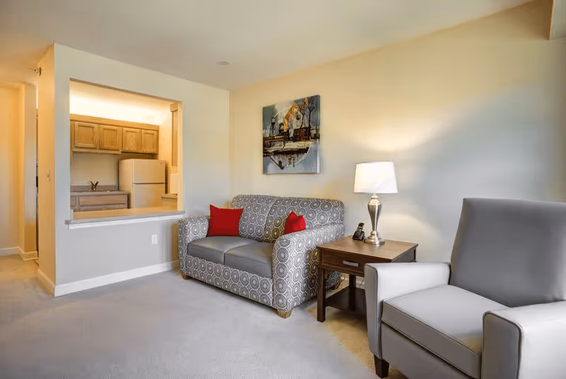A cozy living room area with a patterned loveseat featuring two red pillows, a gray armchair, and a wooden side table with a lamp and a phone. A painting hangs on the wall above the loveseat. The room has beige walls and carpeted floor. In the background, there is a view into a kitchen with wooden cabinets and a refrigerator.