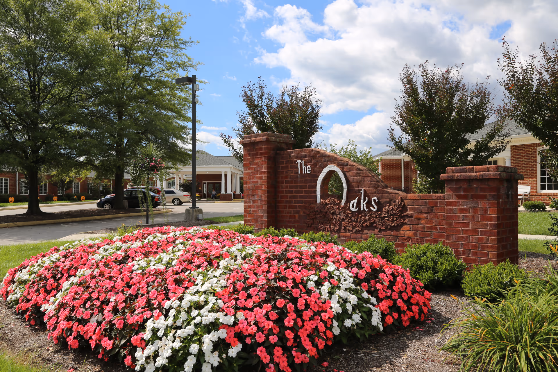 A landscaped entrance area with a brick sign that reads 'The Oaks' surrounded by vibrant pink and white flowers. Behind the sign, there are trees, a parking area with cars, and a building with a covered entrance under a partly cloudy blue sky.