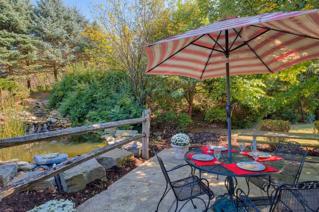Outdoor patio area with a round metal table set for four with plates and glasses, shaded by a large red and white striped umbrella. The patio is next to a small pond with a wooden fence and surrounded by lush green trees and bushes under a clear sky.