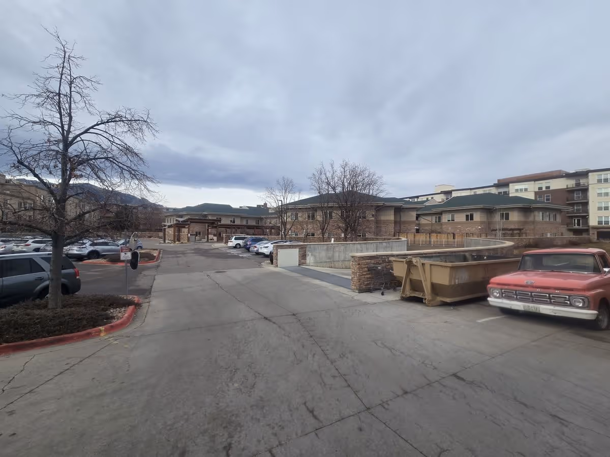 Parking lot and front entrance of a multi-story senior living facility with parked cars and a vintage red pickup under a cloudy sky.