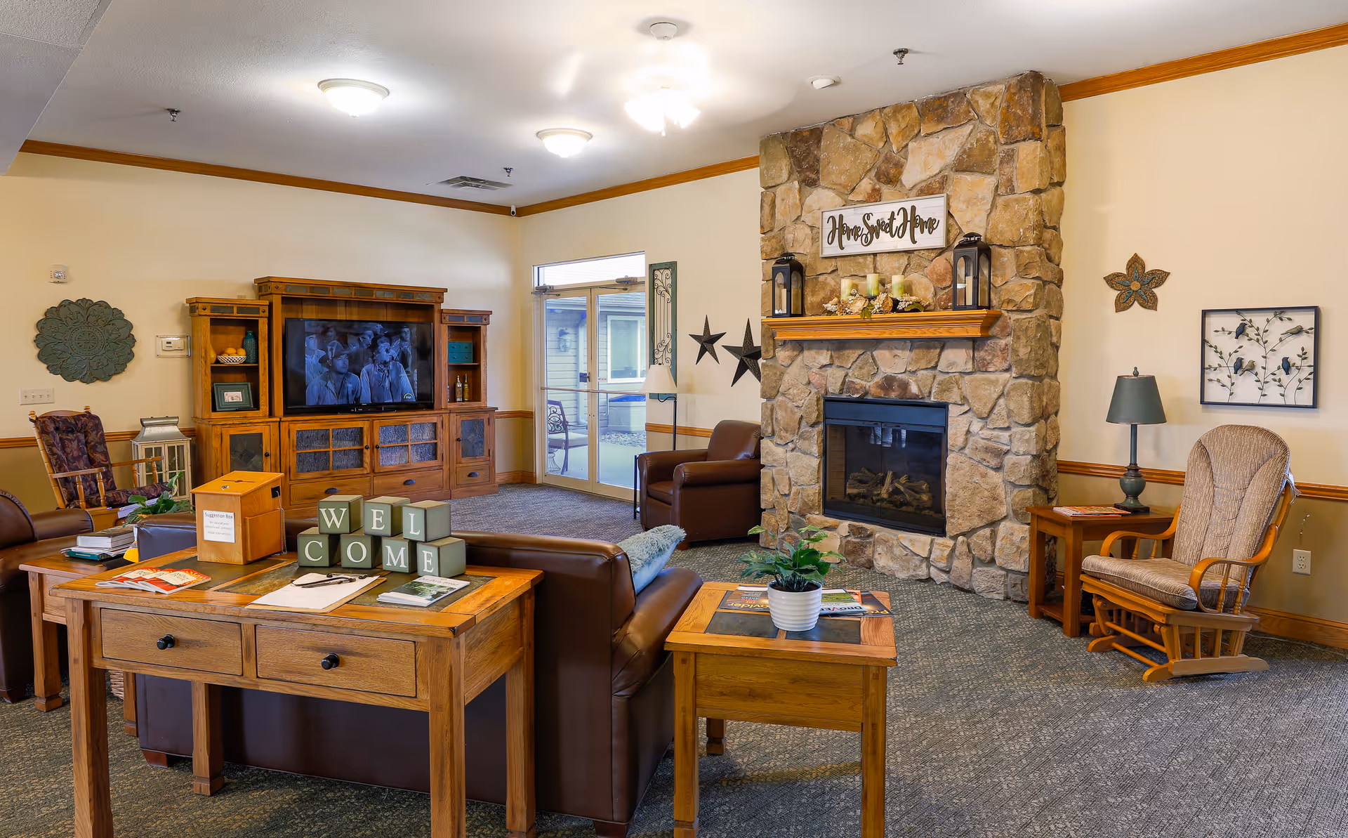 A cozy living room in an assisted living facility featuring a stone fireplace with a 'Home Sweet Home' sign above it. The room includes brown leather sofas, a wooden coffee table with decorative blocks spelling 'WELCOME', a wooden entertainment center with a TV, a rocking chair, and various wall decorations. There is a glass door leading to an outdoor area.