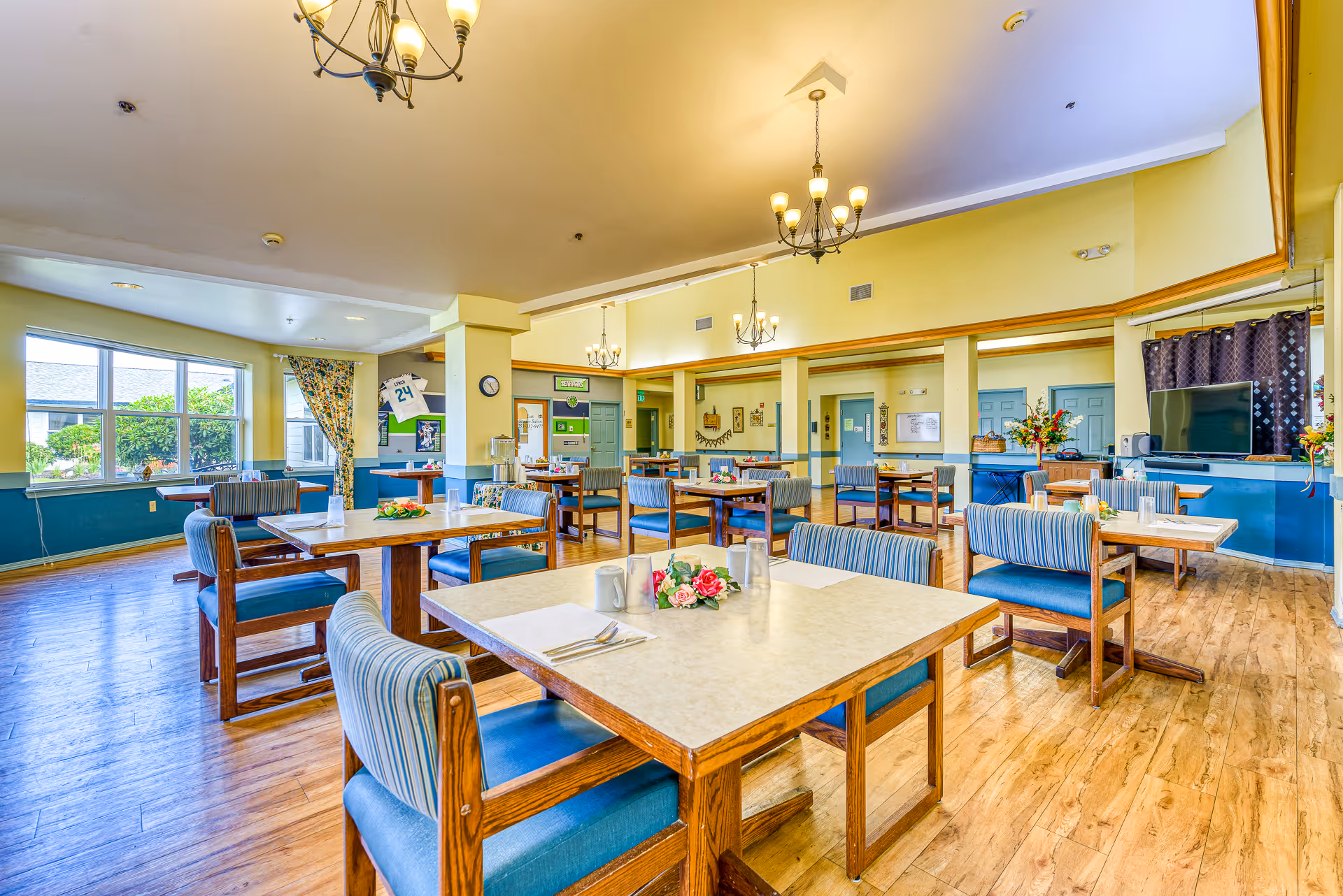 A bright and spacious dining room with multiple wooden tables and blue cushioned chairs. Each table is set with placemats, utensils, cups, and small floral centerpieces. The room has large windows with floral curtains allowing natural light to enter. The walls are painted yellow with blue accents, and there are chandeliers hanging from the ceiling. A television is mounted on the right side of the room with a dark curtain behind it.