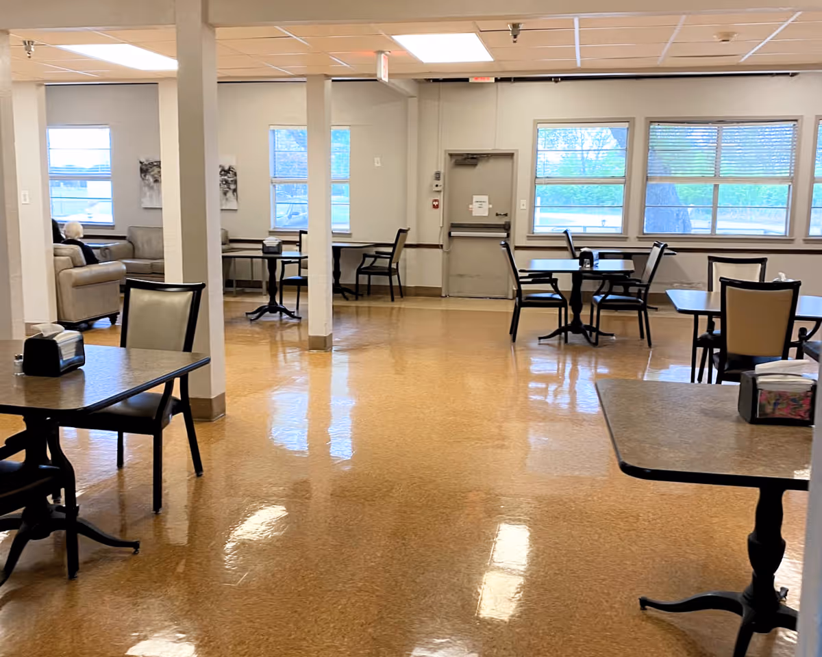 A spacious room with multiple tables and chairs arranged for dining. There are large windows letting in natural light, and a few couches are visible in the background with a person sitting on one of them. The floor is shiny and reflective, and the walls are light-colored with some artwork hanging.