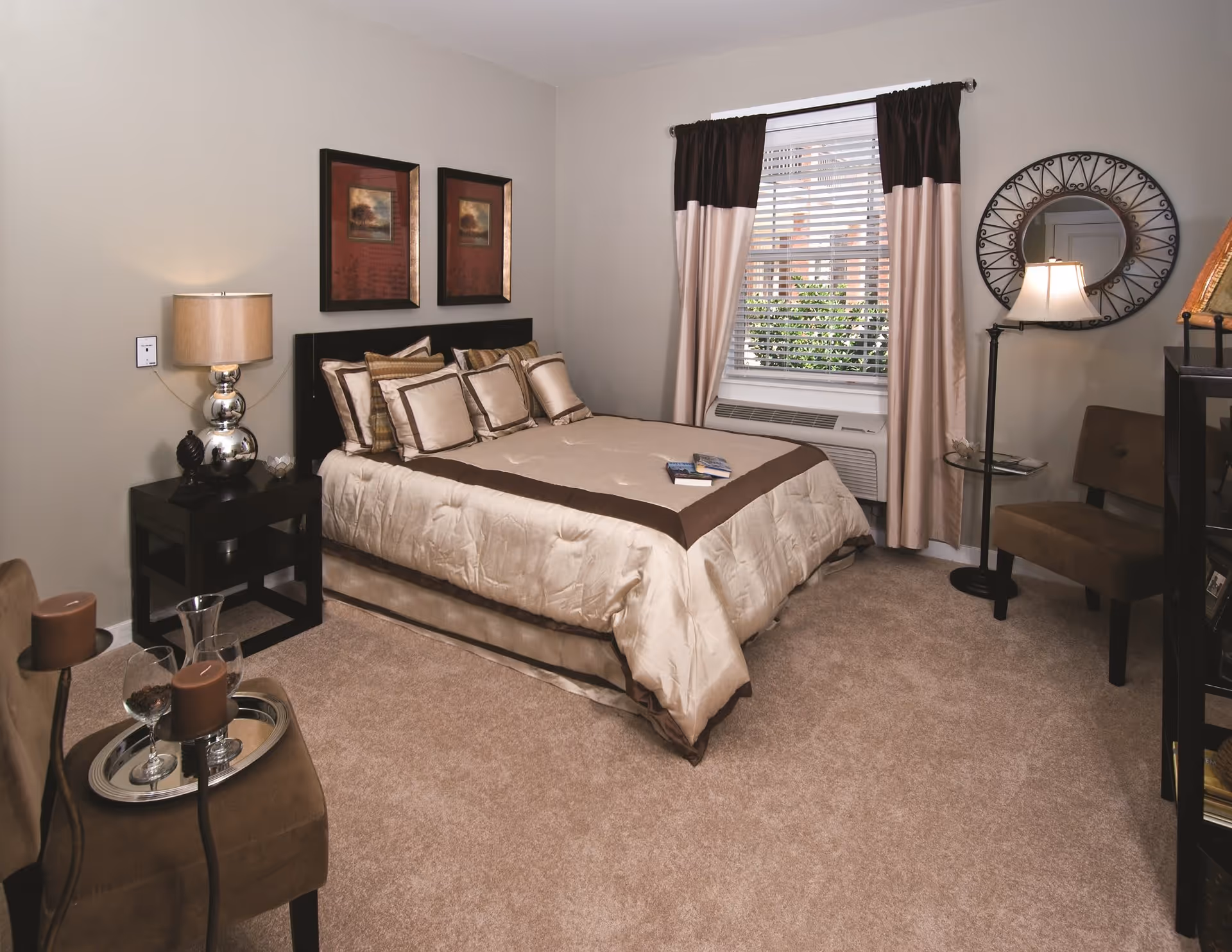 A neatly arranged bedroom with a beige and brown color scheme featuring a bed with multiple pillows, a nightstand with a lamp, two framed pictures on the wall, a window with curtains and blinds, a round decorative mirror, a floor lamp, and two brown chairs with a small table holding candles and glasses.