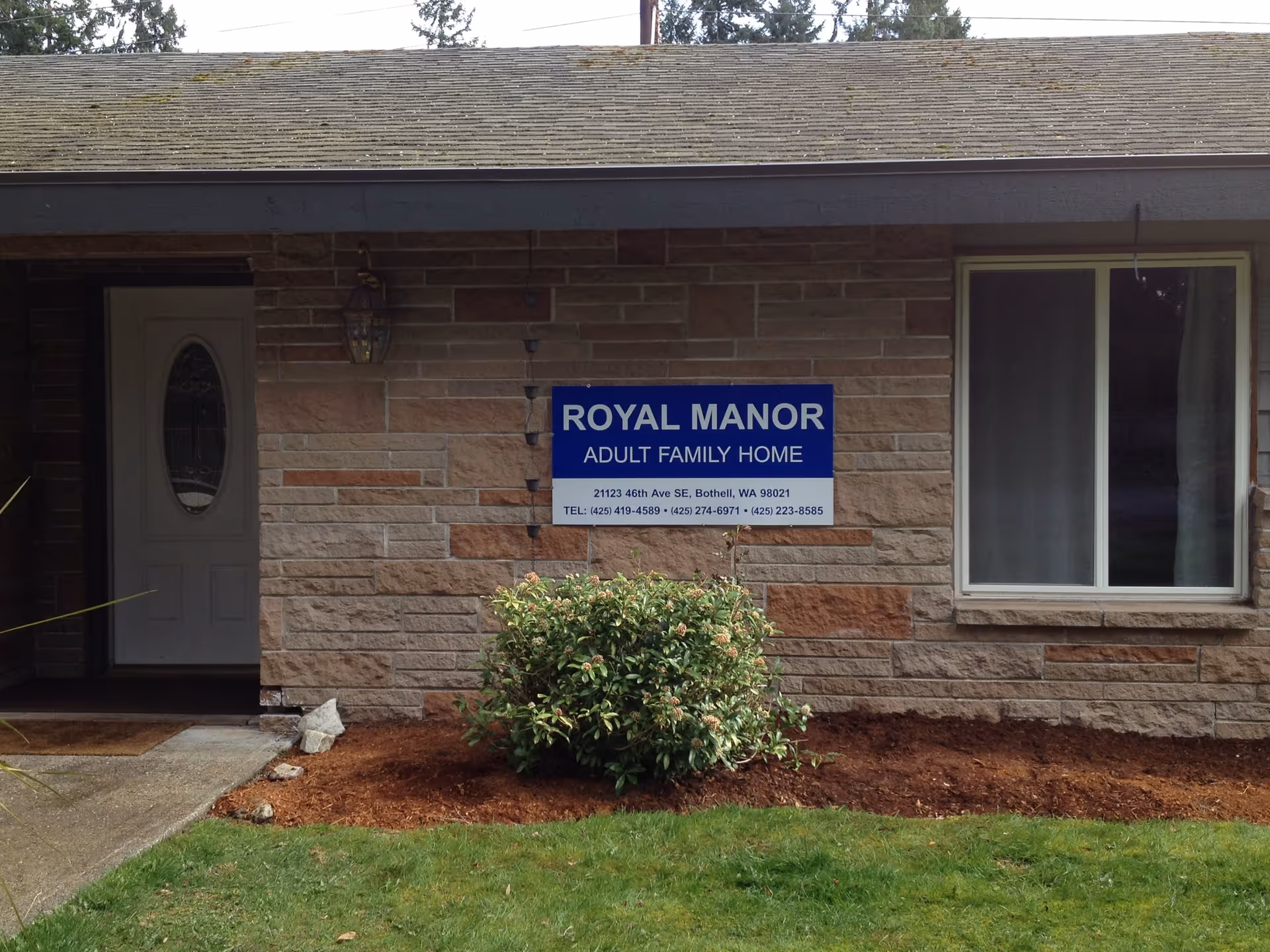 Exterior view of a building wall with a sign that reads 'ROYAL MANOR ADULT FAMILY HOME' along with the address and phone numbers. There is a white door on the left, a window on the right, a small bush in front of the wall, and a grassy area in the foreground.