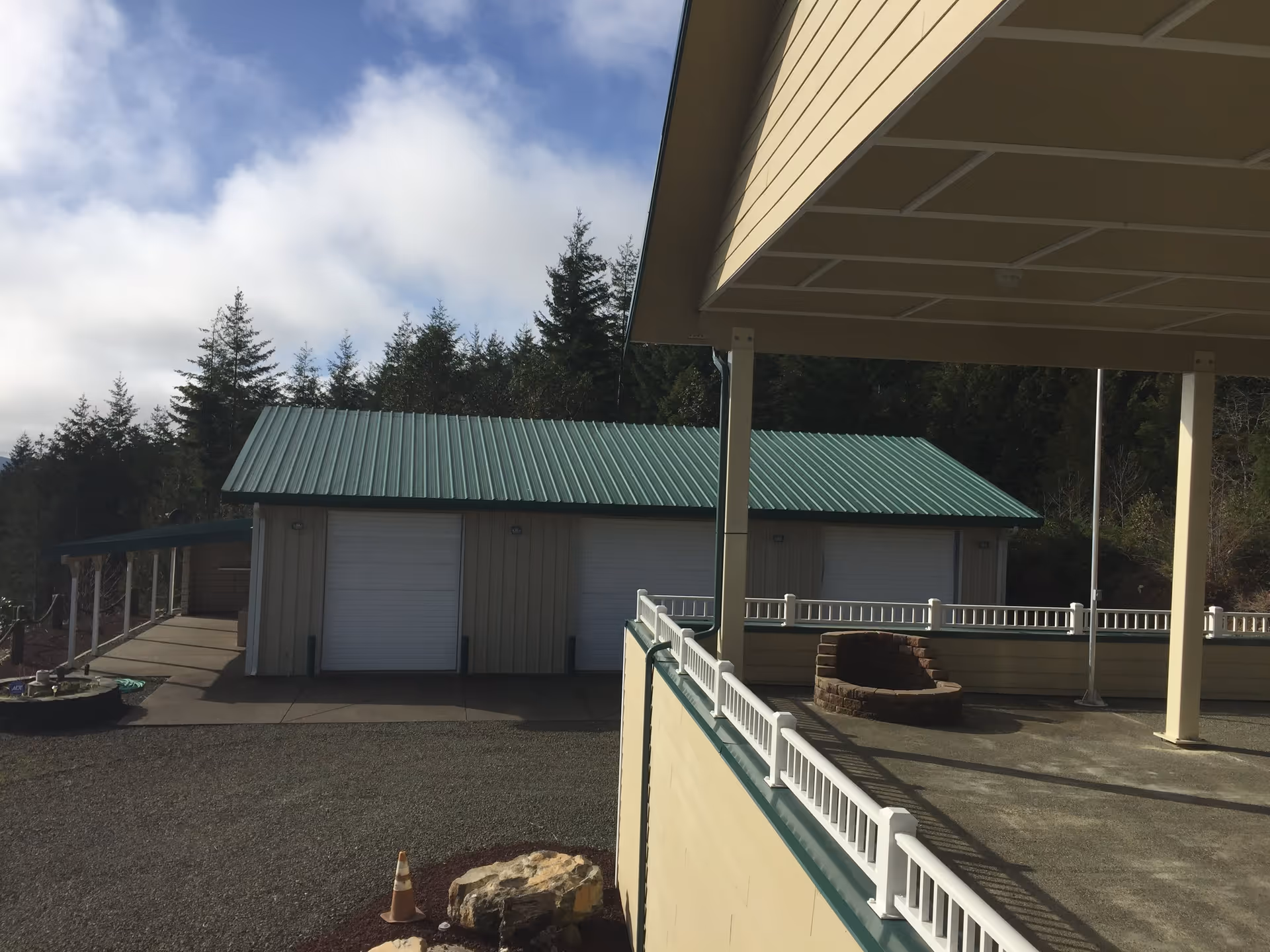 Outdoor view showing a building with a green metal roof and two white garage doors, surrounded by trees and a partly cloudy sky. In the foreground, there is a beige structure with white railings and a small circular brick fire pit.