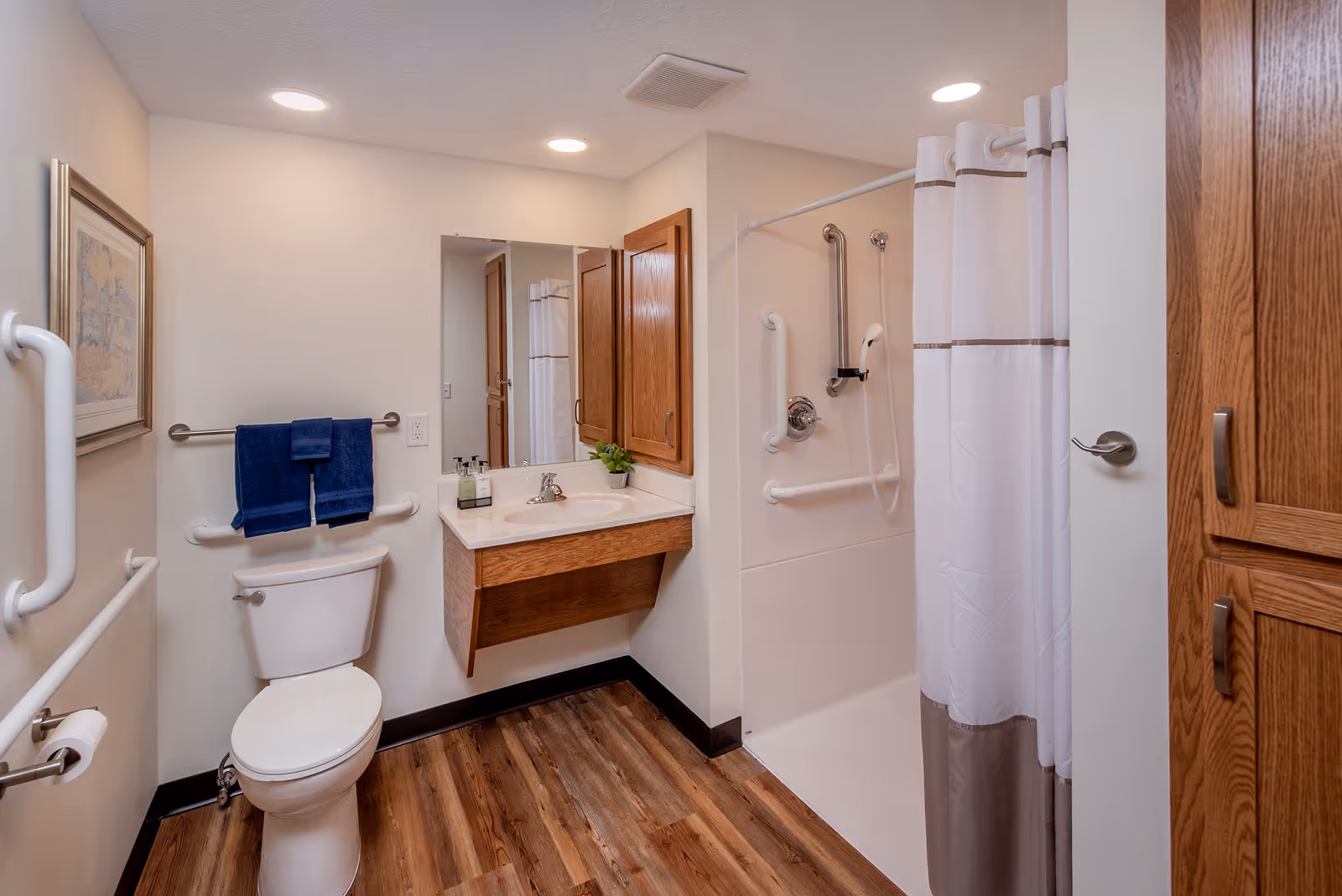 A clean and accessible bathroom with wooden flooring, a white toilet with grab bars, a sink with a mirror above it, a shower area with a white curtain and multiple grab bars, and wooden cabinets on the wall.