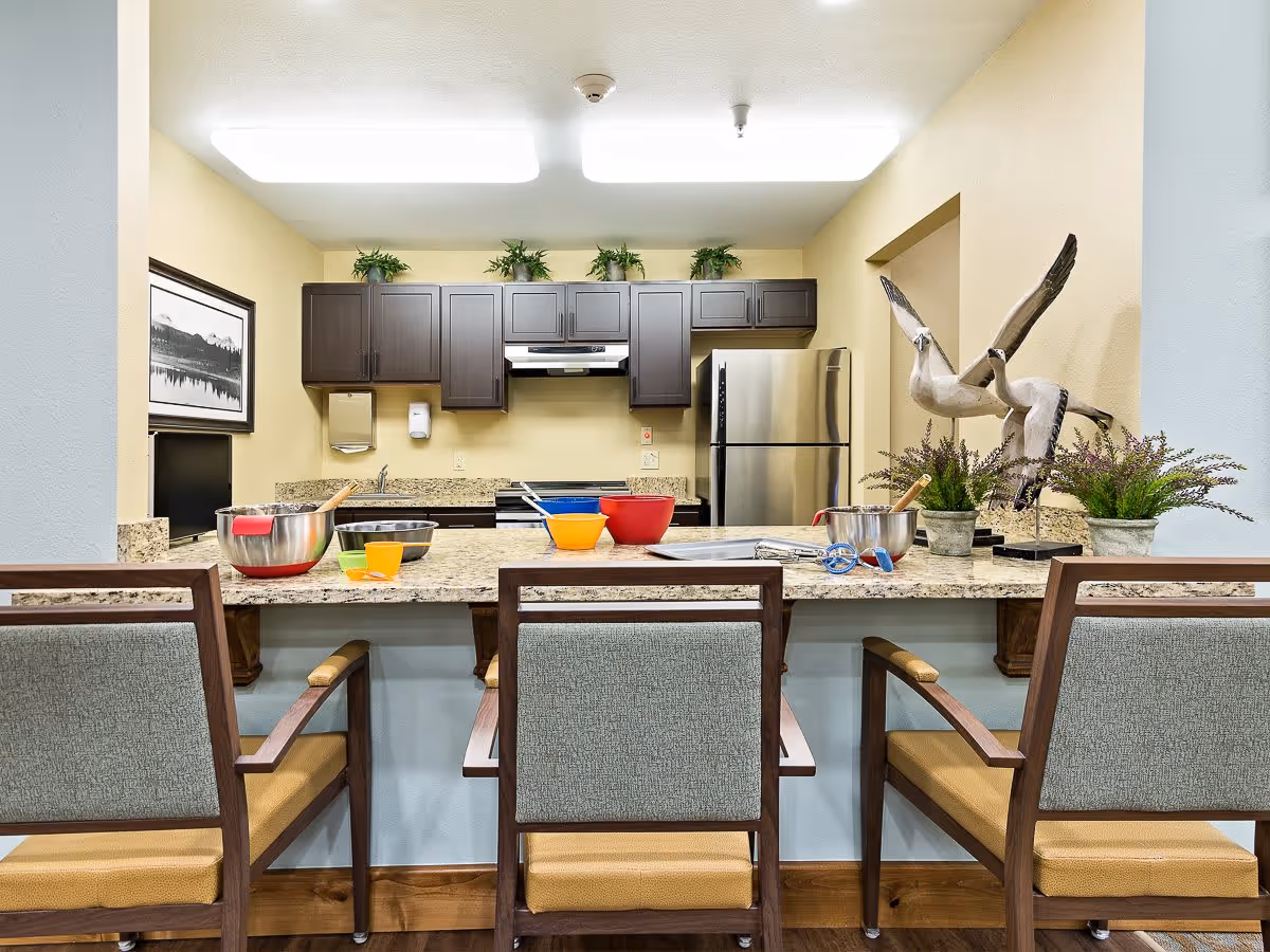 Communal kitchen with a granite countertop, three chairs, bowls and utensils, dark cabinets and a stainless steel refrigerator.