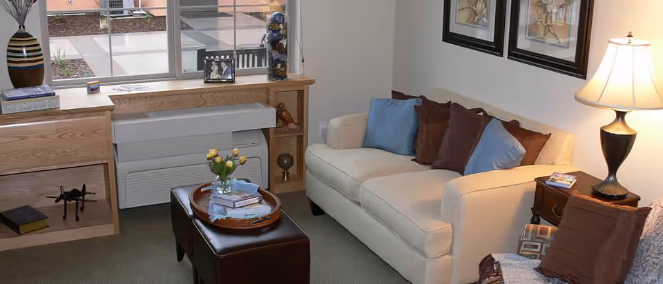 Cozy living room with a beige sofa topped with decorative pillows, an ottoman with a tray, a side table lamp, and built-in shelving under a window.