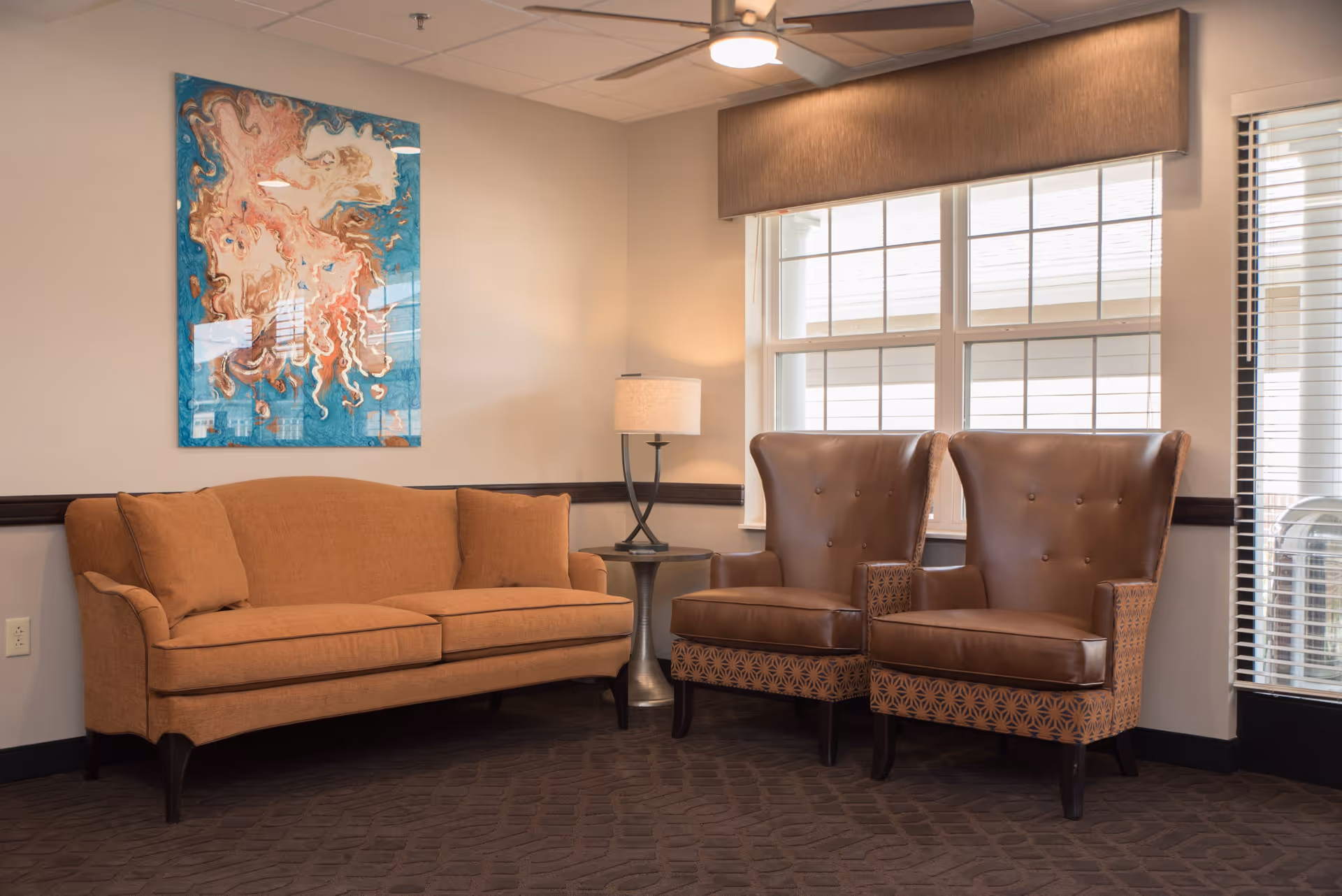 A cozy seating area in a living room with a brown upholstered sofa, two brown leather armchairs with patterned sides, a small round side table with a lamp, and a large window with blinds. A colorful abstract painting hangs on the wall above the sofa.