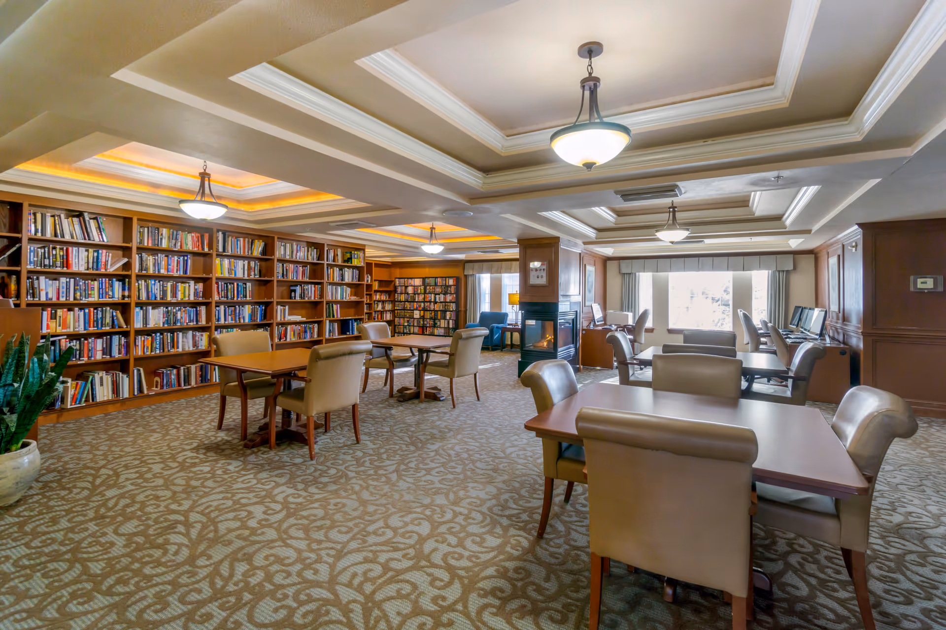 A spacious, well-lit library and common area in a senior living facility with multiple bookshelves filled with books along the left wall, several tables with beige upholstered chairs, a fireplace in the center, and computer stations near a large window on the right side.