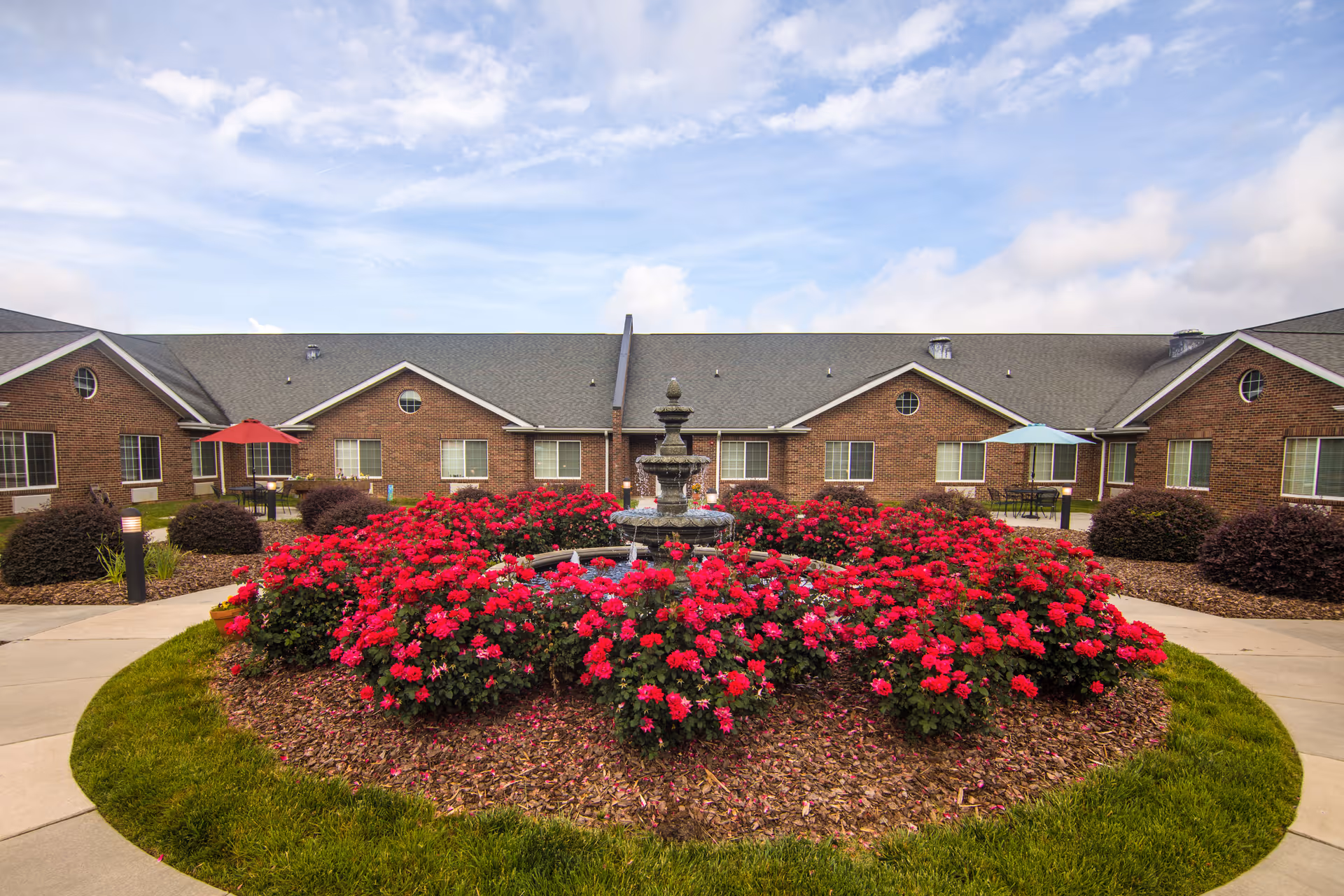Outdoor courtyard area at Camden Health and Rehabilitation featuring a circular flower bed with vibrant red flowers surrounding a multi-tiered water fountain. The courtyard is bordered by a paved walkway and has brick building facades with windows and patio seating with umbrellas in the background under a partly cloudy sky.