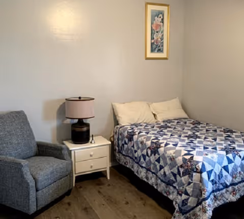 Simple bedroom with a bed covered in a blue patterned quilt, a nightstand with a lamp, and an upholstered chair against a plain wall.