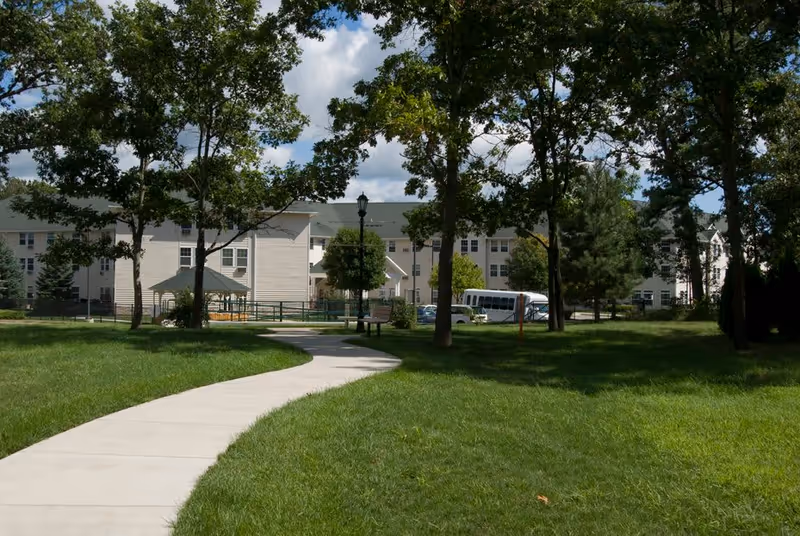 A winding concrete pathway through a grassy area with trees leading to a large multi-story building in the background under a partly cloudy sky. There is a small gazebo and a bench along the path, and a white shuttle bus is parked near the building.