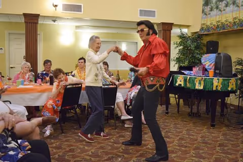 An elderly woman dancing with a man dressed as Elvis Presley in a red shirt and black pants in a senior living community room. Several elderly people are seated around tables covered with orange tablecloths, watching and smiling. The room has warm lighting and a piano decorated with a green 'ALOHA' banner.
