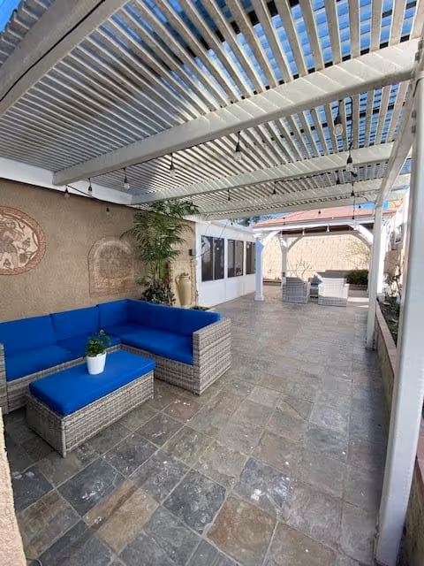 Outdoor covered patio area with a white pergola overhead, featuring a blue cushioned wicker sectional sofa with a matching ottoman and a small potted plant on top. The floor is tiled with stone tiles, and there are additional seating arrangements visible further down the patio. The area is enclosed by walls with some decorative elements and plants.