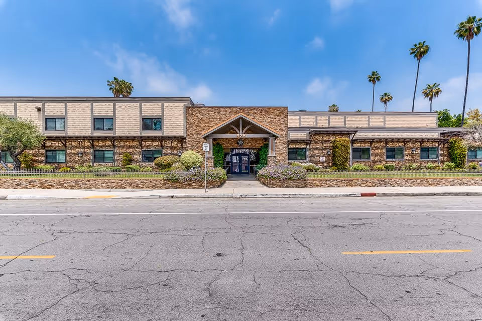 Front exterior view of a single-story senior living facility building with a stone and beige facade, a central entrance with a peaked roof, surrounded by landscaped bushes and palm trees under a blue sky with some clouds.