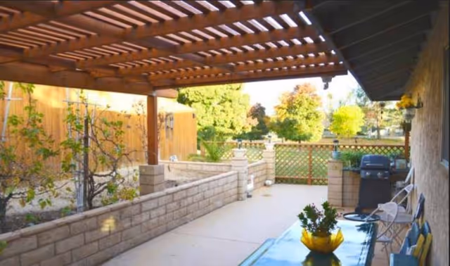 Outdoor patio area with a wooden pergola overhead, stone half-walls, a glass table with a yellow bowl and plant, a barbecue grill, and chairs. The patio overlooks a green lawn with trees and a wooden fence.