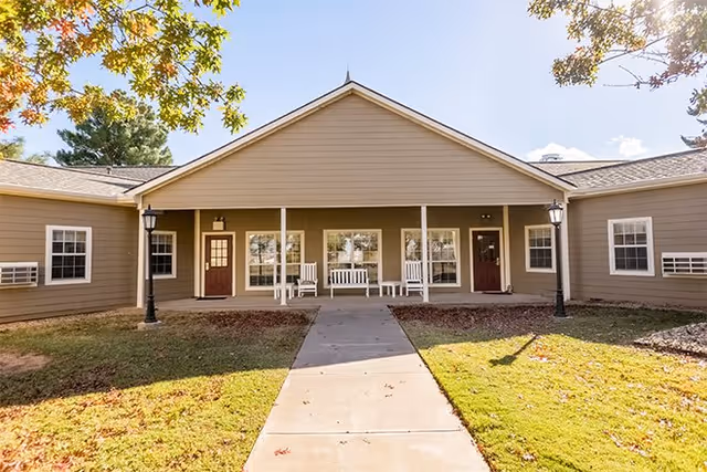 Exterior view of a single-story building with beige siding and a peaked roof. There is a concrete walkway leading to a covered porch area with white rocking chairs and benches. Two lampposts flank the walkway, and trees with autumn leaves frame the scene.