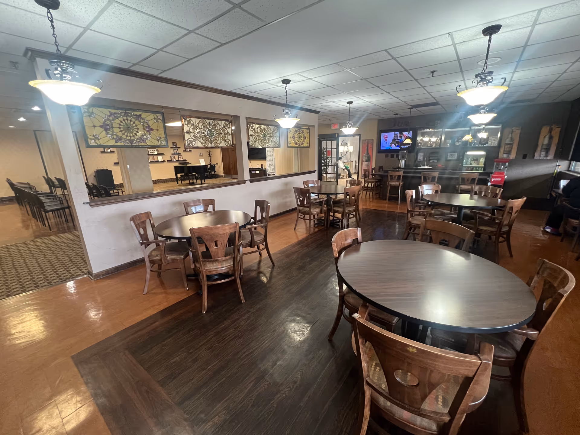 Interior view of a dining area with several round wooden tables and chairs arranged neatly. The room features hanging pendant lights, a large mirror with decorative stained glass panels above it, and a bar area with stools and a television mounted on the wall. The flooring is a mix of wood and carpet, and there is a doorway leading to another room with additional seating.