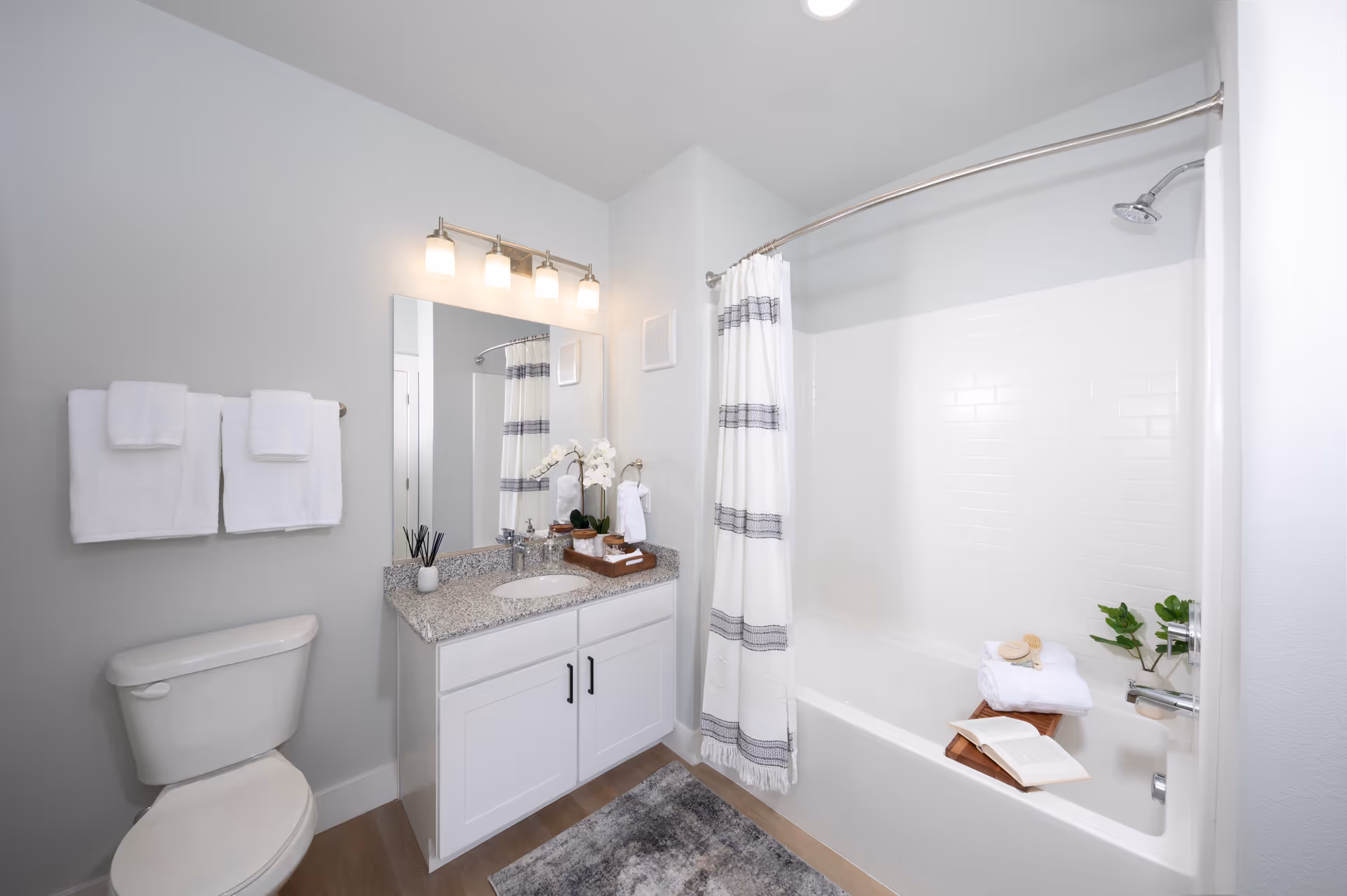 A clean and modern bathroom featuring a white toilet, a vanity with a granite countertop and an oval sink, a large mirror with four light fixtures above it, and a bathtub with a white shower curtain that has gray stripes. The bathtub has a wooden tray holding a rolled towel, a brush, and an open book. There are white towels hanging on a towel rack above the toilet, and a small plant on the bathtub edge. The floor has a gray and beige rug.