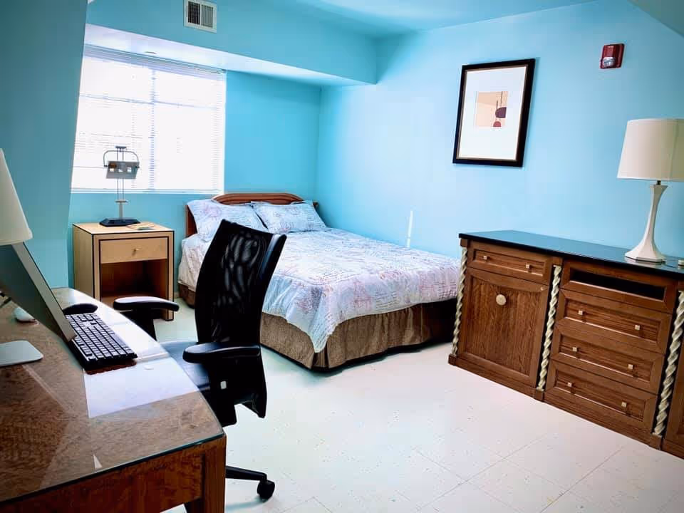 A small bedroom with light blue walls featuring a bed with a patterned quilt, a wooden nightstand with a lamp and a book, a wooden dresser with a lamp on top, a framed abstract artwork on the wall, and a desk with a computer and office chair.