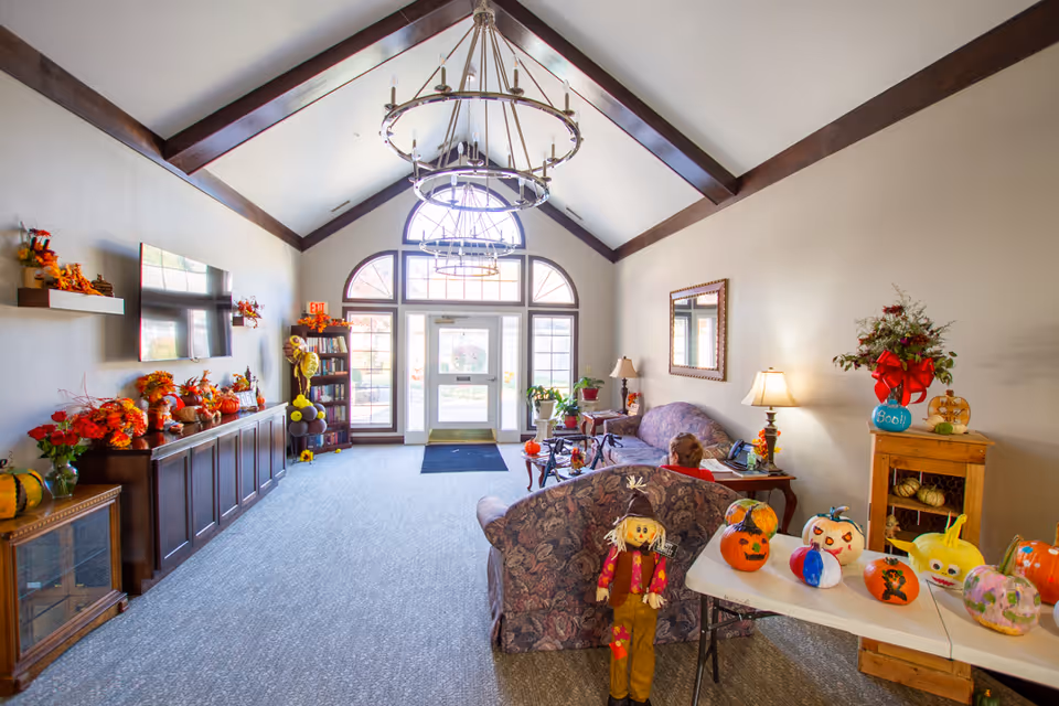 A bright and spacious living room area with high vaulted ceilings and wooden beams. The room is decorated with autumn and Halloween-themed decorations including pumpkins, a scarecrow figure, and fall floral arrangements. There are two patterned sofas, a coffee table, a TV mounted on the wall, and a large windowed door letting in natural light. A person is seated on one of the sofas.